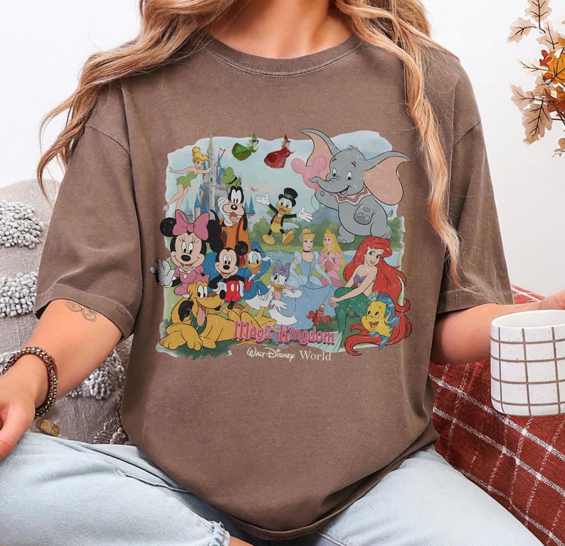 Puede incluir: Camiseta marr&oacute;n de manga corta con un gr&aacute;fico colorido de personajes de Disney, incluyendo Mickey y Minnie Mouse, Donald Duck y Dumbo. El gr&aacute;fico incluye el texto "Magic Kingdom" y "Walt Disney World".