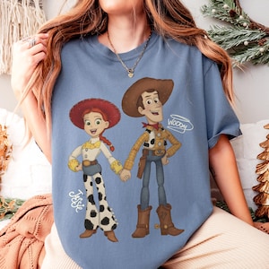 Może przedstawiać: Jasnoniebieska koszulka z kresk&oacute;wkową ilustracją Jessie i Woody'ego z Toy Story. Jessie ma na sobie białą koszulę, spodnie w krowie łaty i czerwony kapelusz. Woody ma na sobie kapelusz kowbojski, ż&oacute;łtą koszulę i niebieskie dżinsy. Na koszulce widnieje napis "Woody".