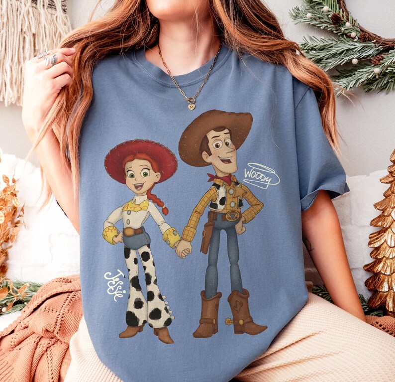 Może przedstawiać: Jasnoniebieska koszulka z kresk&oacute;wkową ilustracją Jessie i Woody'ego z Toy Story. Jessie ma na sobie białą koszulę, spodnie w krowie łaty i czerwony kapelusz. Woody ma na sobie kapelusz kowbojski, ż&oacute;łtą koszulę i niebieskie dżinsy. Na koszulce widnieje napis "Woody".