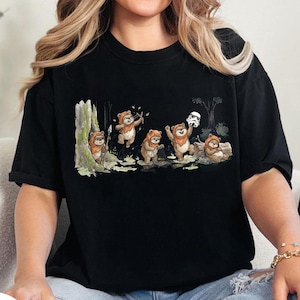 May include: Black t-shirt featuring a whimsical illustration of Ewoks in a forest setting. The design includes several Ewoks, some holding weapons and one with a Stormtrooper helmet. The artwork is detailed and set against a backdrop of trees and foliage.