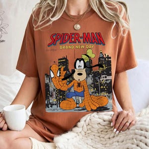 May include: A rust-colored t-shirt featuring a graphic of Spider-Man and Goofy in a cityscape. The text "SPIDER-MAN BRAND NEW DAY" is displayed above the characters. The characters are in a Spider-Man costume.