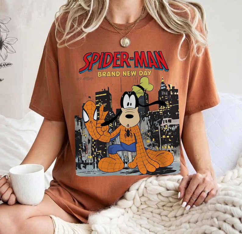 May include: A rust-colored t-shirt featuring a graphic of Spider-Man and Goofy in a cityscape. The text "SPIDER-MAN BRAND NEW DAY" is displayed above the characters. The characters are in a Spider-Man costume.