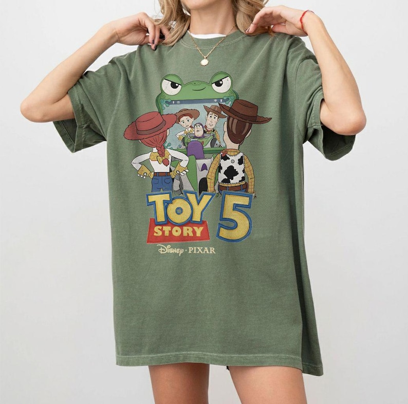 K&ouml;nnte beinhalten: Olivgr&uuml;nes T-Shirt mit einem Grafikdesign aus dem Film Toy Story 5. Das Design zeigt Charaktere wie Woody und Jessie sowie einen Frosch, mit dem Text "TOY STORY 5" in einer bunten Schriftart. Das Disney Pixar-Logo befindet sich unten.
