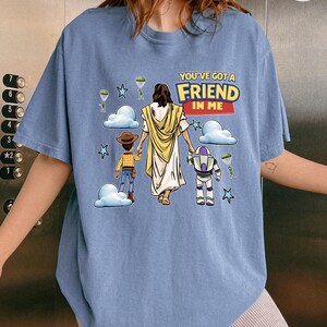 May include: A light blue t-shirt featuring a graphic with the text "YOU'VE GOT A FRIEND IN ME". The graphic depicts Jesus, Woody, and Buzz Lightyear walking together, with clouds, stars, and parachutes in the background.