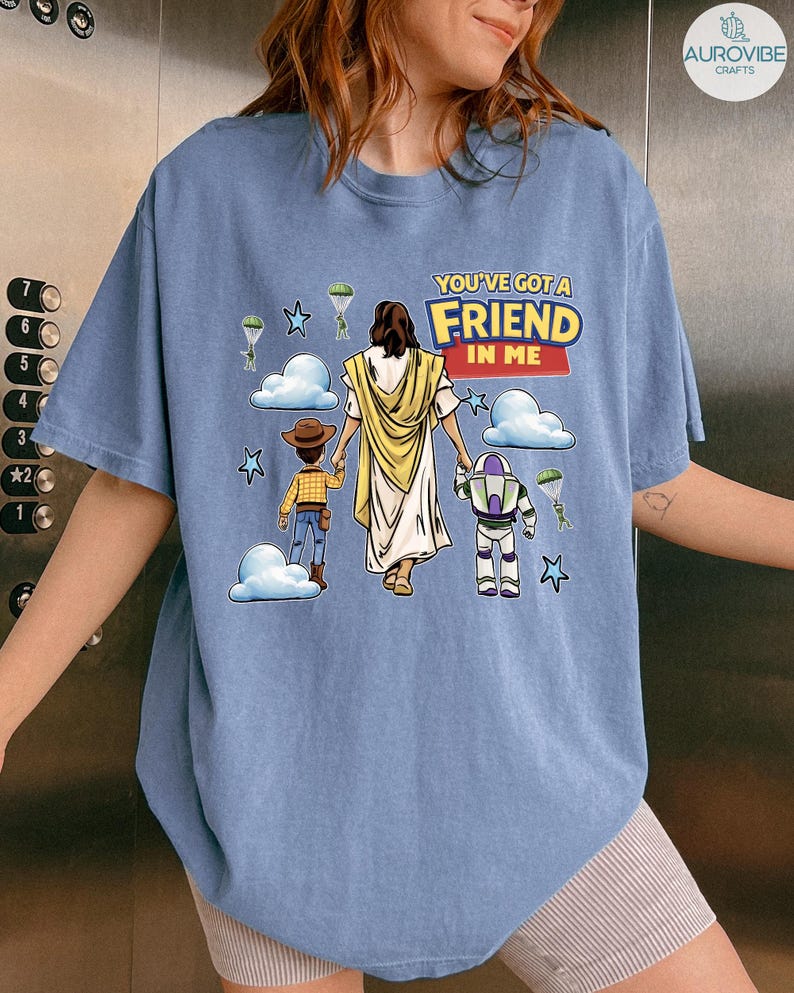 May include: A light blue t-shirt featuring a graphic with the text "YOU'VE GOT A FRIEND IN ME". The graphic depicts Jesus, Woody, and Buzz Lightyear walking together, with clouds, stars, and parachutes in the background.