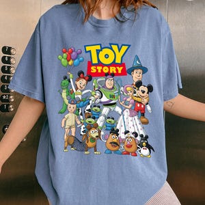 Op de afbeelding: Een lichtblauw T-shirt met de tekst "TOY STORY" in geel en rood, met een kleurrijke afbeelding van personages uit de animatiefilm. Het ontwerp bevat Buzz Lightyear, Woody en andere geliefde personages.