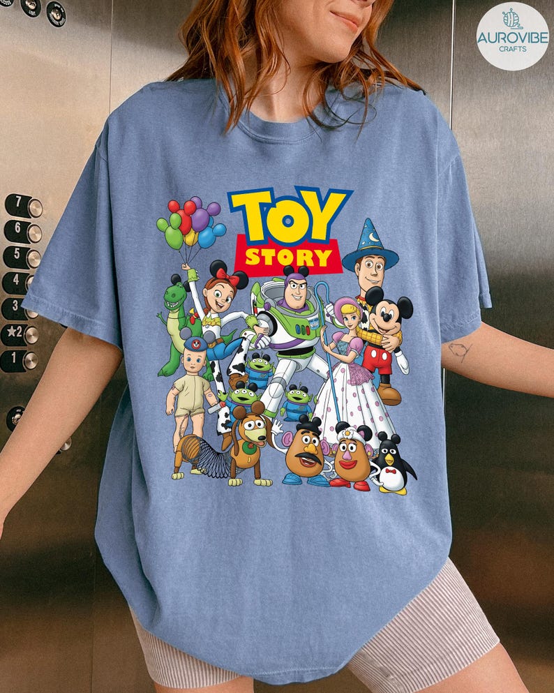Op de afbeelding: Een lichtblauw T-shirt met de tekst "TOY STORY" in geel en rood, met een kleurrijke afbeelding van personages uit de animatiefilm. Het ontwerp bevat Buzz Lightyear, Woody en andere geliefde personages.