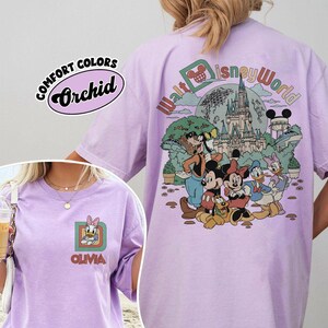 May include: Orchid-colored t-shirt with a Disney World graphic. The front features a Daisy Duck design with the name "Olivia." The back displays Disney characters and the words "Walt Disney World." The shirt is labeled "Comfort Colors."