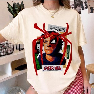 Op de afbeelding: Cr&egrave;mekleurig T-shirt met een Spider-Man-afbeelding. Het ontwerp bevat een portret van Spider-Man met de tekst "SPIDER-MAN BRAND NEW DAY" en de woorden "WITH GREAT POWER COMES GREAT RESDONSBILITY" en "DAILY BUGLE".