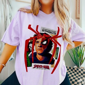 Op de afbeelding: Lichtpaars T-shirt met een Spider-Man-afbeelding. Het ontwerp bevat een portret van het personage, een rood spinnenweb en de tekst "SPIDER-MAN BRAND NEW DAY" en "WITH GREAT POWER COMES GREAT RESPONSIBILITY."