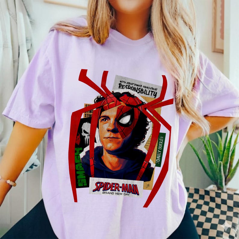 Op de afbeelding: Lichtpaars T-shirt met een Spider-Man-afbeelding. Het ontwerp bevat een portret van het personage, een rood spinnenweb en de tekst "SPIDER-MAN BRAND NEW DAY" en "WITH GREAT POWER COMES GREAT RESPONSIBILITY."