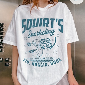 May include: White t-shirt with a teal graphic featuring a cartoon turtle wearing a snorkel mask. The text reads "Squirt's Snorkeling" above the turtle and "Fin, Noggin, Dude" below, with "-East Australian Current-" in between.