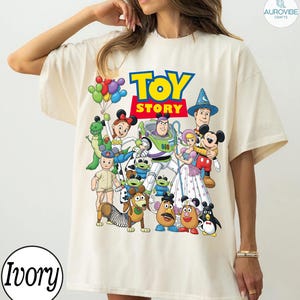 Op de afbeelding: Ivoor kleurig T-shirt met een kleurrijke afbeelding van Toy Story-personages, waaronder Buzz Lightyear en Woody. De woorden "TOY STORY" staan in grote, vette letters. Het shirt heeft een ronde hals en korte mouwen. Het woord "Ivory" staat linksonder.