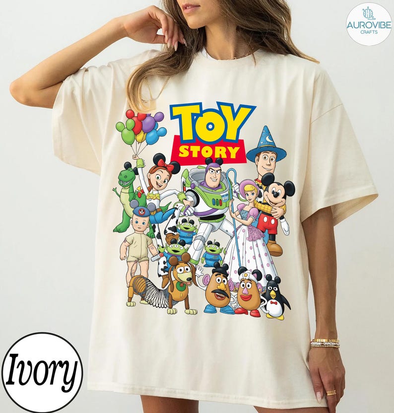Op de afbeelding: Ivoor kleurig T-shirt met een kleurrijke afbeelding van Toy Story-personages, waaronder Buzz Lightyear en Woody. De woorden "TOY STORY" staan in grote, vette letters. Het shirt heeft een ronde hals en korte mouwen. Het woord "Ivory" staat linksonder.