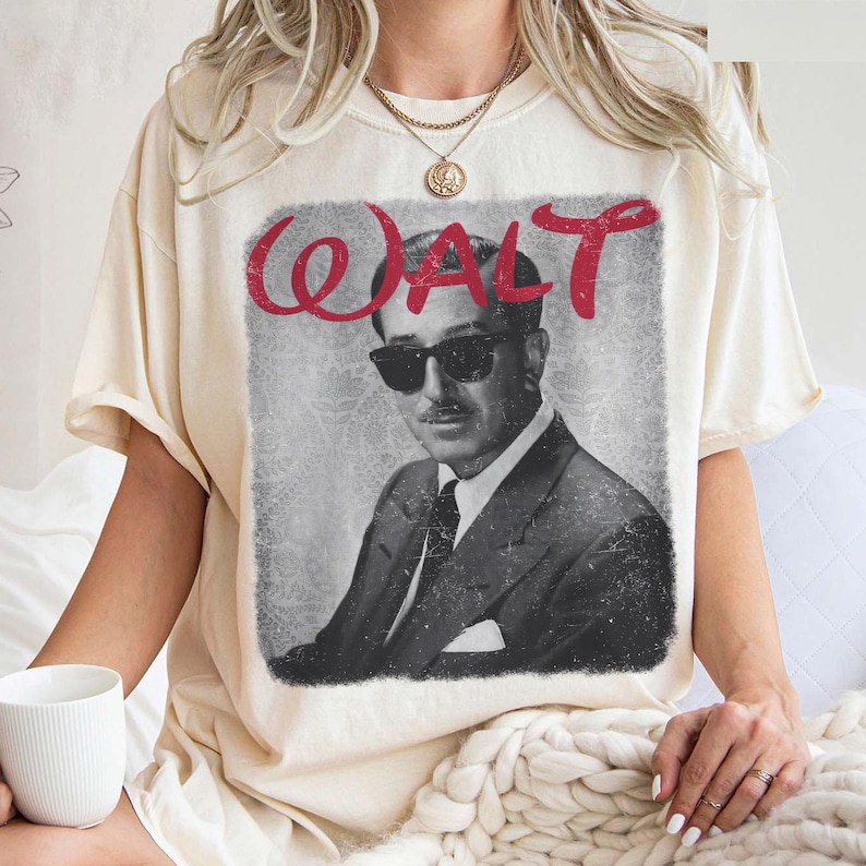 May include: Cream-colored t-shirt featuring a black and white image of a man in sunglasses and a suit, with the name "WALT" in red script above the image. The shirt has short sleeves and a crew neck.