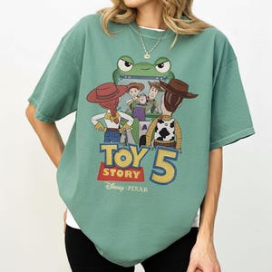 K&ouml;nnte beinhalten: Gr&uuml;nes Kurzarm-T-Shirt mit einem Grafikdesign aus dem Film Toy Story 5. Das Design zeigt Charaktere wie Woody und Buzz Lightyear. Das Shirt hat auch das Disney Pixar-Logo.