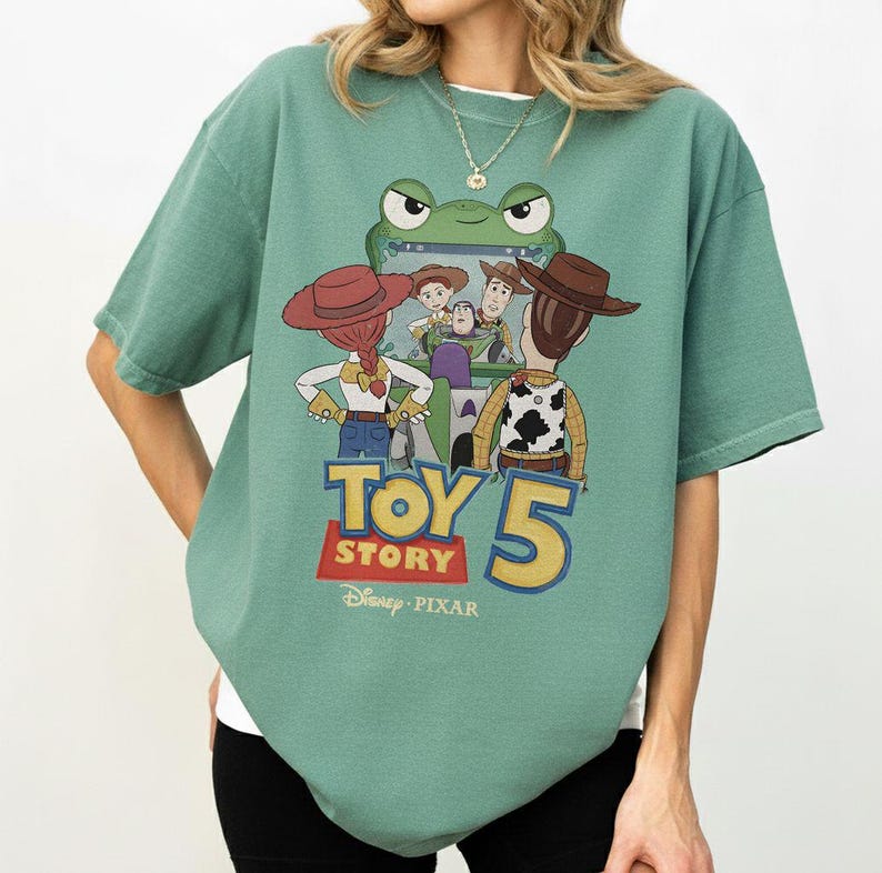 K&ouml;nnte beinhalten: Gr&uuml;nes Kurzarm-T-Shirt mit einem Grafikdesign aus dem Film Toy Story 5. Das Design zeigt Charaktere wie Woody und Buzz Lightyear. Das Shirt hat auch das Disney Pixar-Logo.