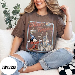May include: Espresso-colored t-shirt featuring a graphic of Mickey Mouse in a wizard outfit, the words "Magic Kingdom", and a castle. The shirt is a short-sleeved, crew neck style.