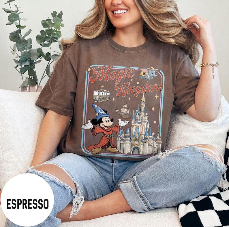 May include: Espresso-colored t-shirt featuring a graphic of Mickey Mouse in a wizard outfit, the words "Magic Kingdom", and a castle. The shirt is a short-sleeved, crew neck style.