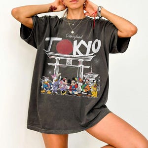 Puede incluir: Camiseta gris oscuro con un dise&ntilde;o de Disneyland Tokyo. El dise&ntilde;o incluye un c&iacute;rculo rojo, texto blanco, una puerta japonesa y personajes de Disney, como Mickey y Minnie Mouse, Donald Duck y Goofy.