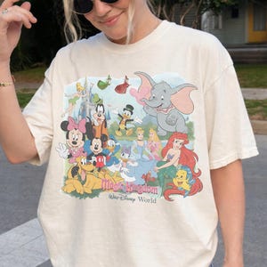 Puede incluir: Camiseta color crema con un gr&aacute;fico colorido de personajes de Disney, incluyendo Mickey y Minnie Mouse, Donald Duck y Dumbo. El gr&aacute;fico incluye el texto "Magic Kingdom Walt Disney World".