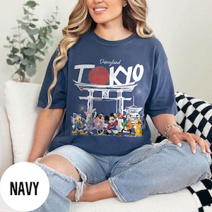 Puede incluir: Camiseta azul marino con un dise&ntilde;o de Disneyland Tokyo. El dise&ntilde;o presenta un sol rojo, una puerta japonesa y la palabra "Tokyo" en blanco. La camiseta muestra personajes de dibujos animados. Un estilo casual.