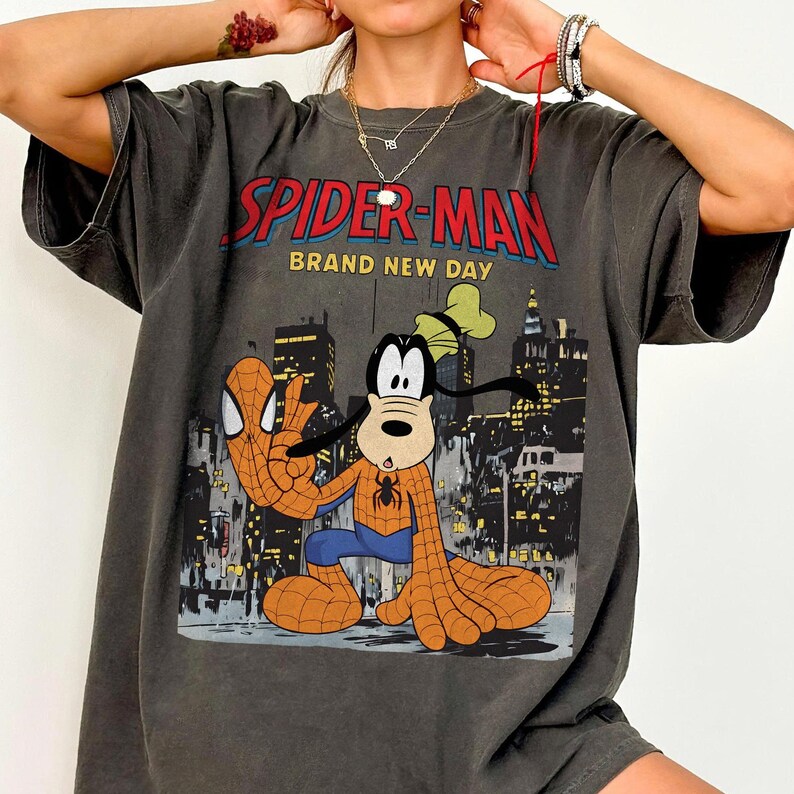 May include: Dark gray t-shirt featuring a graphic of Spider-Man and Goofy in a city setting. The text "SPIDER-MAN BRAND NEW DAY" is printed across the top in red and yellow. The shirt has short sleeves and a relaxed fit.