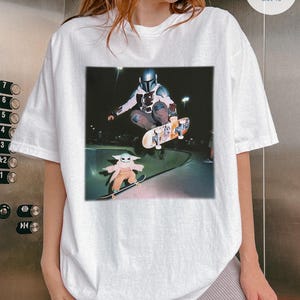 Puede incluir: Camiseta blanca con un gr&aacute;fico de un personaje con casco haciendo monopat&iacute;n, y una imagen m&aacute;s peque&ntilde;a de un personaje beb&eacute; en monopat&iacute;n. La imagen est&aacute; sobre un fondo oscuro, con un entorno de parque de patinaje.