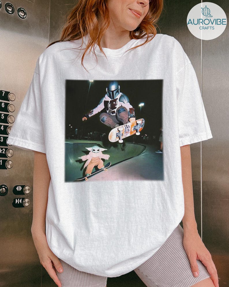 Puede incluir: Camiseta blanca con un gr&aacute;fico de un personaje con casco haciendo monopat&iacute;n, y una imagen m&aacute;s peque&ntilde;a de un personaje beb&eacute; en monopat&iacute;n. La imagen est&aacute; sobre un fondo oscuro, con un entorno de parque de patinaje.