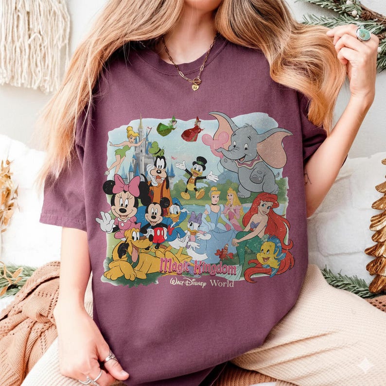 Puede incluir: Una camiseta granate con un gr&aacute;fico colorido de personajes de Disney, incluyendo Mickey y Minnie Mouse, Dumbo y Ariel. El gr&aacute;fico incluye el texto "Magic Kingdom Walt Disney World".