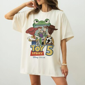 K&ouml;nnte beinhalten: Cremefarbenes, &uuml;bergro&szlig;es T-Shirt mit einem Grafikdesign aus dem Film "Toy Story 5". Das Design zeigt Charaktere wie Woody und Buzz Lightyear, mit dem Filmtitel in fetten Buchstaben. Das Disney Pixar-Logo ist ebenfalls zu sehen.