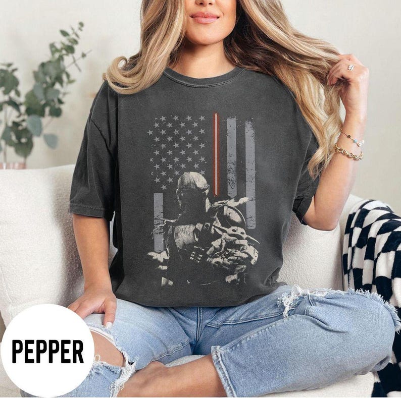 May include: Dark gray t-shirt featuring a distressed American flag design with a red stripe and a silhouette of a character holding a baby. The word "PEPPER" is printed at the bottom.