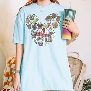 May include: A light blue t-shirt featuring a graphic print of various snacks and treats arranged in the shape of a mouse head silhouette. A banner across the center contains the text "BEST DAY EVER!" surrounded by small stars and food icons.