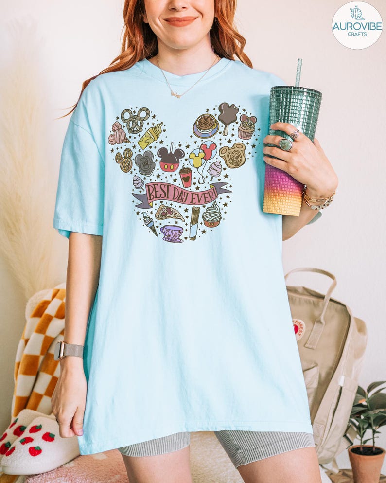 May include: A light blue t-shirt featuring a graphic print of various snacks and treats arranged in the shape of a mouse head silhouette. A banner across the center contains the text "BEST DAY EVER!" surrounded by small stars and food icons.
