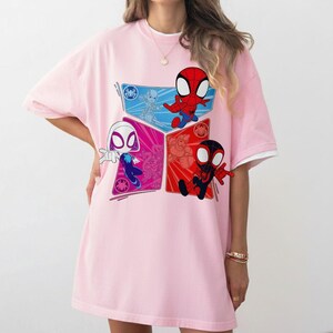 May include: A light pink t-shirt featuring a colorful graphic of Spider-Man characters. The design includes Spider-Man, Spider-Gwen, and Miles Morales in a cartoon style. The shirt has short sleeves with white trim.