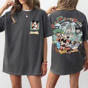 May include: Dark gray t-shirt featuring Mickey Mouse on the front and a Walt Disney World graphic on the back. The front design includes Mickey Mouse in a square frame with the name "JAMES" below. The back design shows Disney characters and a castle.
