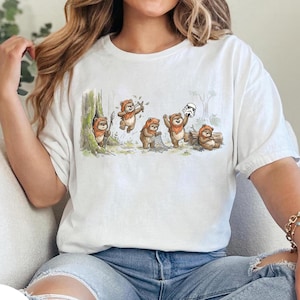 May include: White t-shirt featuring a whimsical illustration of Ewoks in a forest setting. The cartoon depicts several Ewoks engaged in various activities, including playing instruments and holding a net, with a Stormtrooper helmet nearby.