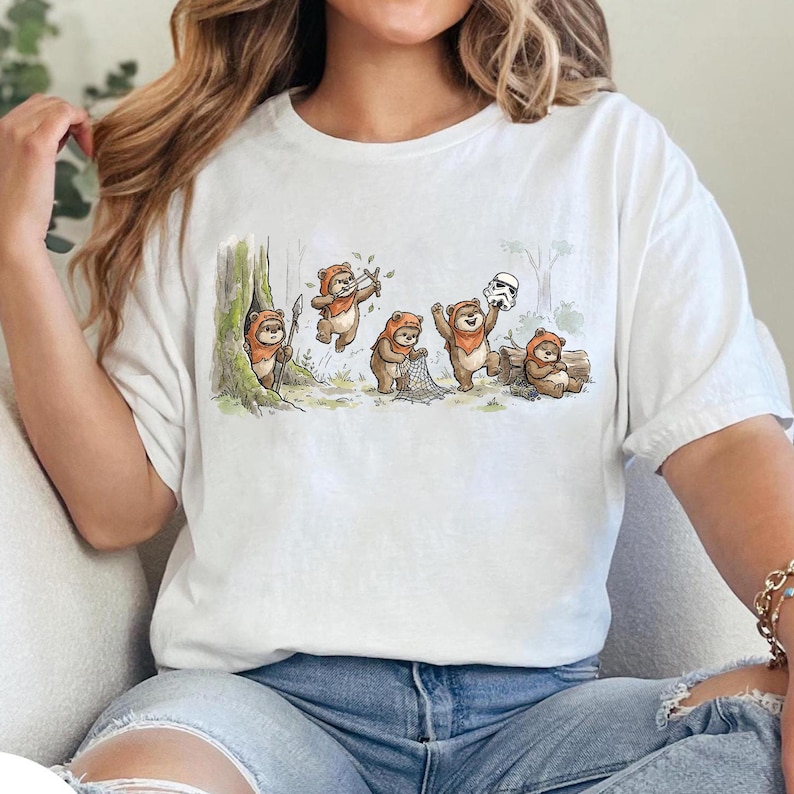 May include: White t-shirt featuring a whimsical illustration of Ewoks in a forest setting. The cartoon depicts several Ewoks engaged in various activities, including playing instruments and holding a net, with a Stormtrooper helmet nearby.