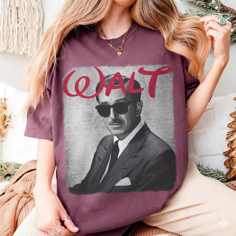 May include: Burgundy-colored t-shirt featuring a black and white image of a man wearing sunglasses and a suit. The word "WALT" is printed in red cursive above the image. The shirt is made of a soft material.