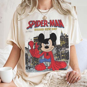 May include: Cream-colored t-shirt featuring a graphic of Spider-Man and Mickey Mouse in a comic book style. The text "SPIDER-MAN BRAND NEW DAY" is printed across the top. The design includes a cityscape background.