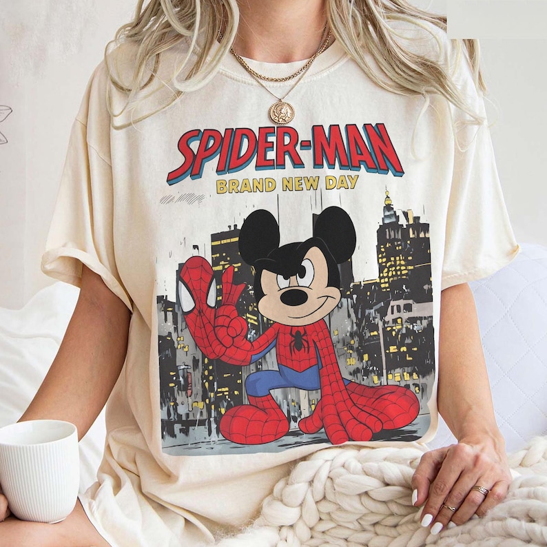 May include: Cream-colored t-shirt featuring a graphic of Spider-Man and Mickey Mouse in a comic book style. The text "SPIDER-MAN BRAND NEW DAY" is printed across the top. The design includes a cityscape background.