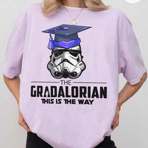 Pu&ograve; includere: Una maglietta lilla con un elmetto da Stormtrooper con un cappello di laurea. Il testo recita "THE GRADALORIAN THIS IS THE WAY" in nero. Il disegno &egrave; centrato sulla maglietta.