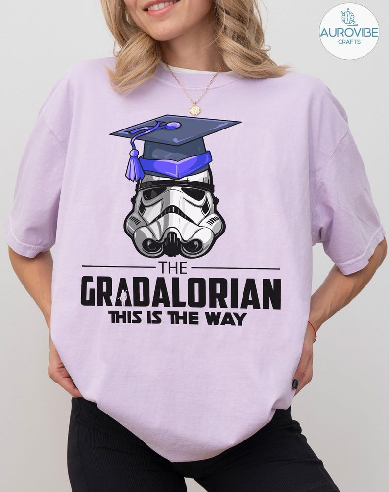 Pu&ograve; includere: Una maglietta lilla con un elmetto da Stormtrooper con un cappello di laurea. Il testo recita "THE GRADALORIAN THIS IS THE WAY" in nero. Il disegno &egrave; centrato sulla maglietta.