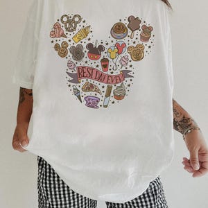May include: A white t-shirt featuring a graphic print of various food items arranged in the shape of a mouse head silhouette. The design includes a banner with the text BEST DAY EVER! surrounded by small stars and colorful snacks and drinks.