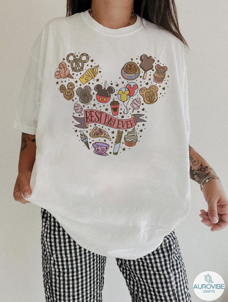 May include: A white t-shirt featuring a graphic print of various food items arranged in the shape of a mouse head silhouette. The design includes a banner with the text BEST DAY EVER! surrounded by small stars and colorful snacks and drinks.
