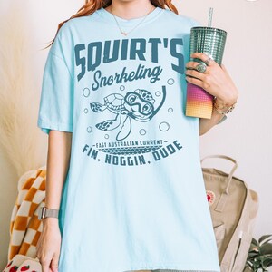 May include: Light blue t-shirt with the text "Squirt's Snorkeling" and a cartoon turtle wearing goggles. The shirt also features the phrase "East Australian Current - Fin, Noggin, Dude."