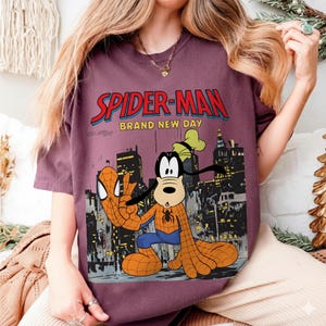 May include: A maroon t-shirt featuring a graphic of Spider-Man and Goofy in a cityscape. The text "SPIDER-MAN BRAND NEW DAY" is displayed in red and blue. Goofy is dressed as Spider-Man, with a spider emblem on his chest.