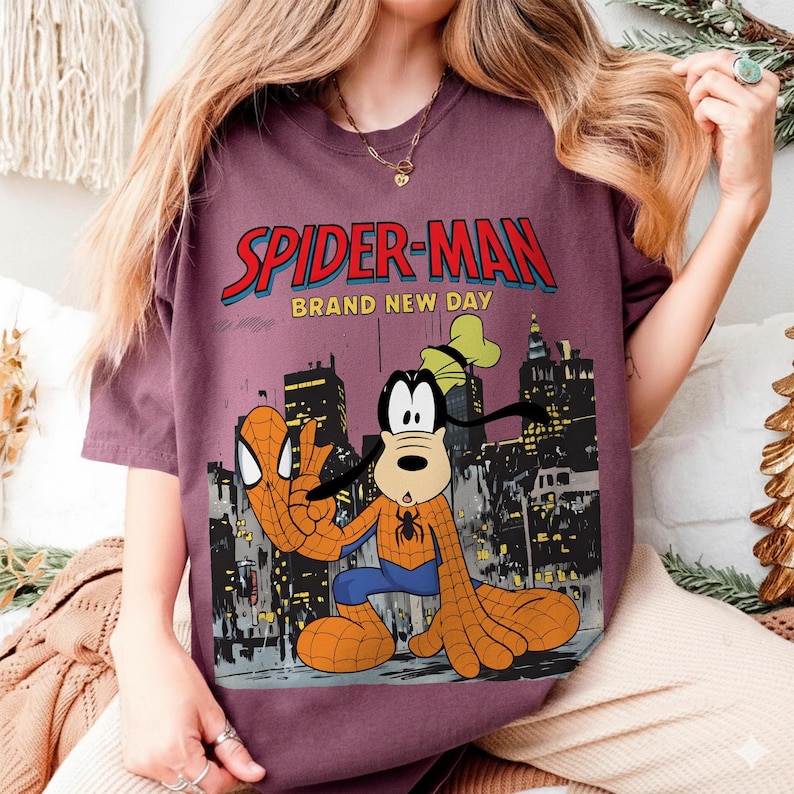 May include: A maroon t-shirt featuring a graphic of Spider-Man and Goofy in a cityscape. The text "SPIDER-MAN BRAND NEW DAY" is displayed in red and blue. Goofy is dressed as Spider-Man, with a spider emblem on his chest.