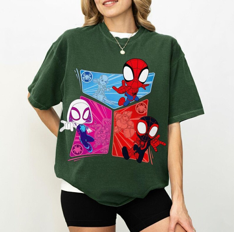 May include: Dark green t-shirt featuring a colorful graphic of Spider-Man characters. The design includes Spider-Man, Spider-Gwen, and Miles Morales in a comic book style. The shirt has a crew neck and short sleeves.
