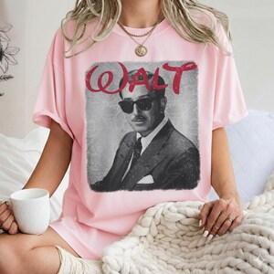 May include: Pink t-shirt featuring a black and white image of a man in a suit and sunglasses, with the name "WALT" in red script above the image. The shirt has short sleeves and a crew neck.